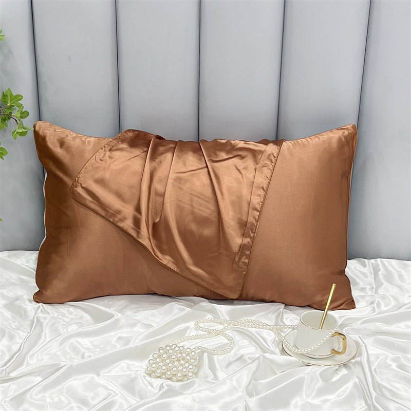 An Aloha Relax Cooling Beauty Sleep Pillowcase in brown, made of luxurious double-sided ice silk, rests on a white satin sheet with a matching blanket. Nearby, a teacup with straw and pearl necklace add charm. The gray upholstered headboard and small plant complete the serene scene.