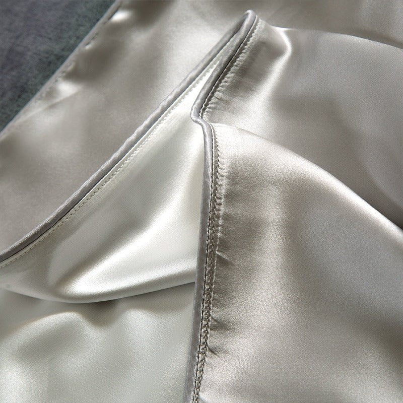 Close-up of an Aloha Relax Cooling Beauty Sleep Pillowcase in shiny silver, made from double-sided ice silk. The smooth, reflective fabric has visible stitching along the edge and elegantly lies on a contrasting surface, perfect for hair and skin protection for hot sleepers.