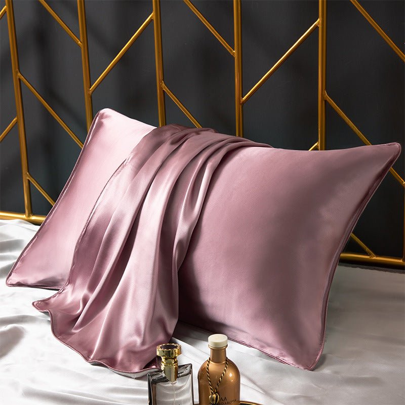 A silky Cooling Beauty Sleep Pillowcase by Aloha Relax rests against a gold geometric headboard, complemented by a matching pink silk scarf draped over it. In the foreground, two perfume bottles sit elegantly on a pristine white surface.