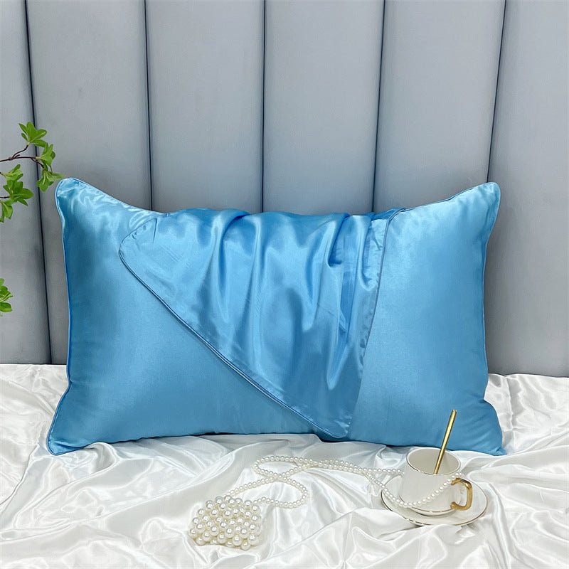 A blue Cooling Beauty Sleep Pillowcase by Aloha Relax lies on a white sheet in front of a tufted gray headboard. Green foliage appears to the left, while a white teacup with a gold spoon sits beside pearls in the foreground.