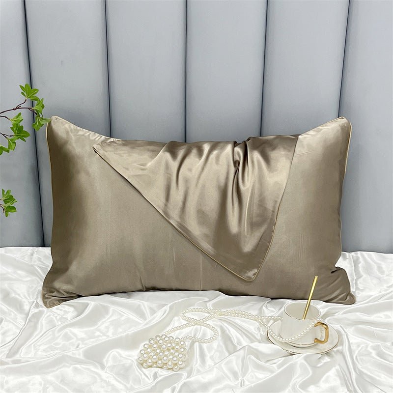 An Aloha Relax Cooling Beauty Sleep Pillowcase in golden silk graces a bed with a white satin sheet. Nearby are a pearl necklace, gold-spooned cup, and green leaves. A light gray padded headboard adds understated charm to the elegant ambiance.