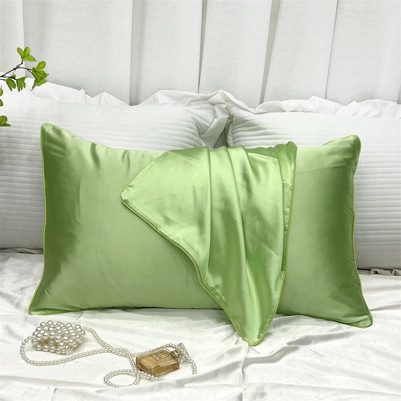 An Aloha Relax Cooling Beauty Sleep Pillowcase in green silk rests on a white bed with a matching silk blanket. Elegantly placed to the left are pearls and a small wrapped soap. A leafy branch peeks from the top corner, enhancing the scene's charm with reactive printing.