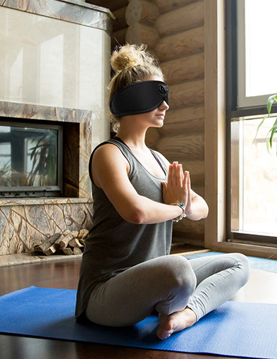 A person sits cross-legged on a blue yoga mat near a window, wearing a grey tank top, grey leggings, and the Aloha Relax 3D Bluetooth Sleep Mask With Breathable Intelligence. Hands are in a prayer position, and a wooden wall and fireplace are visible in the background.