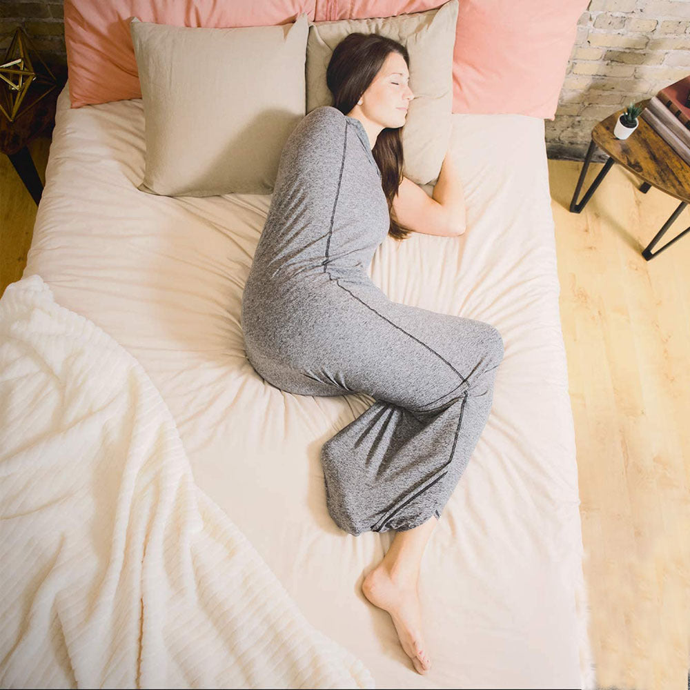 A woman with long brown hair, dressed in a gray long-sleeve top and matching gray pants, is lying on her side in an Aloha Relax Comfortable Sleep Pod (for Parent or Child) on a bed with light-colored sheets and pillows. To the left of the bed sits a light-colored blanket, while to the right stands a small table holding an alarm clock.