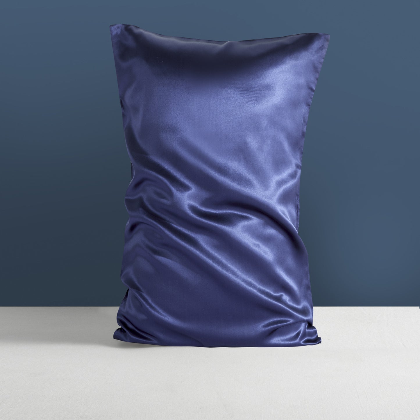 A dark blue, double-sided silk sleep pillowcase from Aloha Relax's "Silk Silk Heavy" collection is elegantly positioned against a blue-gray backdrop. The smooth, mulberry silk fabric catches the light, highlighting its luxurious texture and reflecting a sense of opulence as the pillow stands on a light-hued surface.