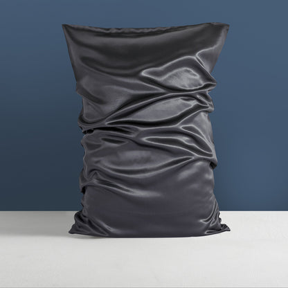 An Aloha Relax Double-sided Silk Sleep Pillowcase, in an elegant black color, is vertically positioned against a blue wall. Its shiny fabric, reminiscent of mulberry silk, displays distinct wrinkles and folds. The pillowcase rests on a light-colored surface.