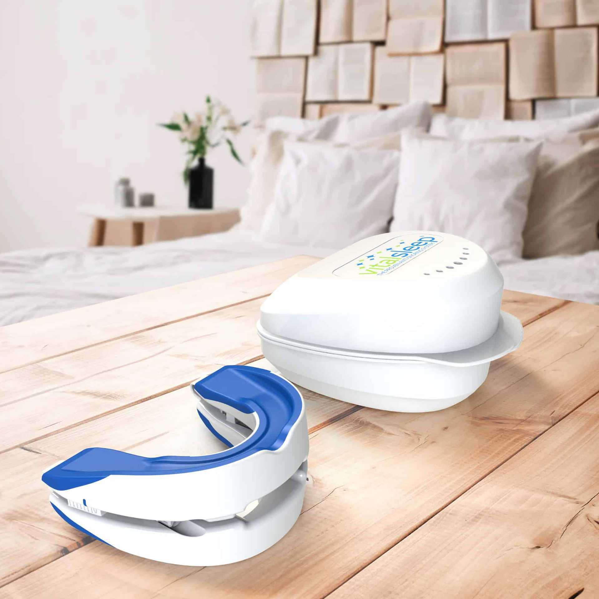 Two white and blue Anti-Snoring Mouthpiece Devices by Aloha Relax rest on a wooden table. One is open, displaying the adjustable mouth guard, while the other is closed with a digital display. In the background, there's a neatly made bed with white pillows and a wall decorated with open books, evoking an atmosphere of sleep improvement.