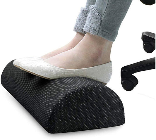 A person wearing grey pants and white shoes with a textured pattern is resting their feet on the Aloha Relax Foot Rest Relaxing Body Pad (Adjustable), which features a dotted surface for stress relief. The semi-cylindrical black footrest is placed on the floor next to a chair with visible black wheels.