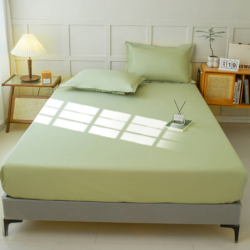 A neatly made bed featuring the Aloha Relax Waterproof Mattress Protector, surrounded by light green fitted sheets and pillows, is in a well-lit room. On one side, a small bedside table with a lamp and calendar stands, while a wooden shelf adorned with decor items occupies the other side. Sunlight casts its window pattern across the solid color fabric of the bed.