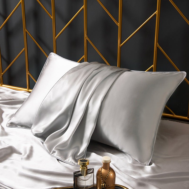 A bed is adorned with an Aloha Relax Cooling Beauty Sleep Pillowcase in light silver, enhancing a luxurious geometric-patterned gold headboard. Skincare bottles rest elegantly on the sheet, complementing the refined setting achieved through advanced reactive printing techniques.