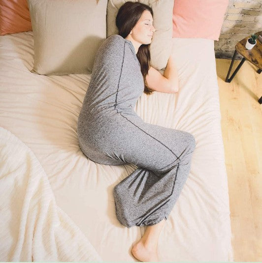 A person with long hair lies on a bed, sleeping on their side in a gray, full-length sleeved sleeping gown. The bed, featuring the Comfortable Sleep Pod by Aloha Relax, is equipped with multiple pillows and has a light-colored duvet designed for stress relief. A small wooden side table is visible beside the bed.