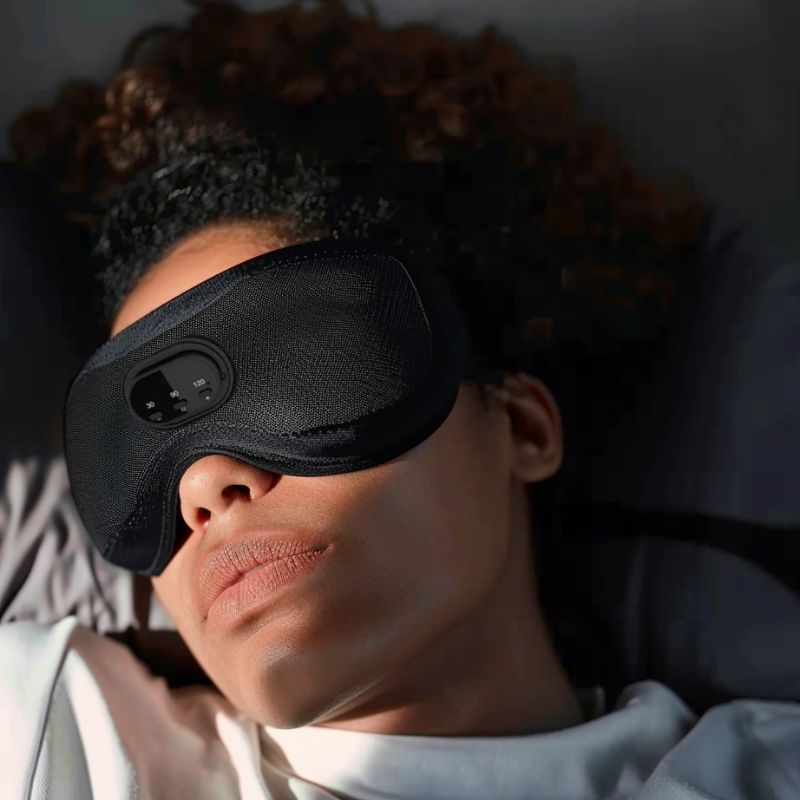 A person with curly hair is lying down, wearing an Aloha Relax 3D Bluetooth Sleep Mask With Breathable Intelligence, which has electronic components on the front. The person appears relaxed, resting on a gray pillow and wearing a white shirt. The room lighting is dim, creating the perfect ambiance for restful sleep during travel.